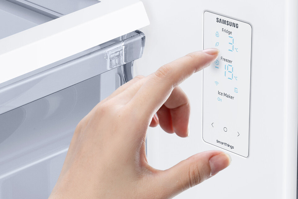 Osoba dotyka panelu sterowania lodówki z wyświetlaczem pokazującym temperatury: Fridge 3°C, Freezer -18°C. Na panelu widoczne są napisy: \'SAMSUNG\', \'Ice Maker On\', \'smartthings\'.