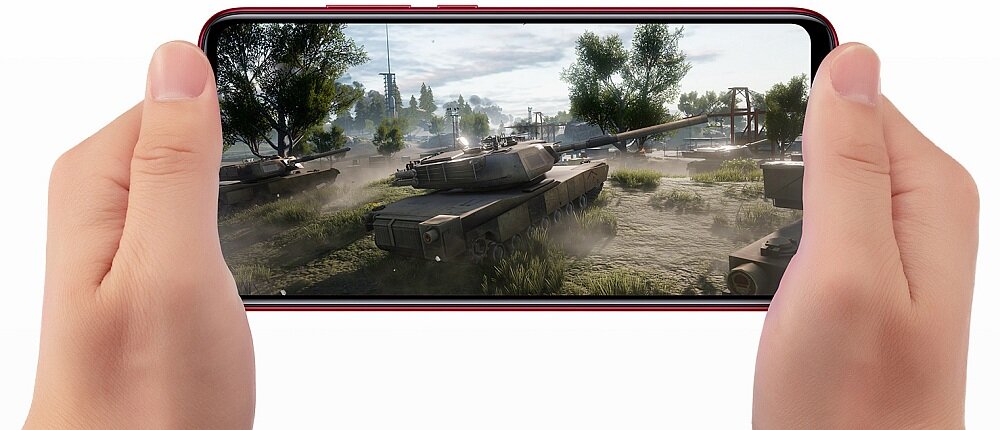 Osoba trzyma smartfon, na ekranie którego wyświetlana jest gra z czołgami i drzewami w tle. Obudowa telefonu jest czerwona.