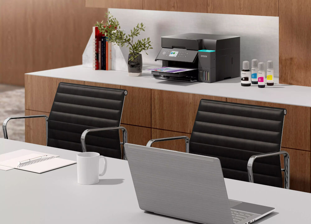 Na biurku znajduje się urządzenie wielofunkcyjne z logo EPSON, obok którego stoją cztery butelki z tuszem. W tle widać drewnianą szafkę z książkami i rośliną w wazonie.