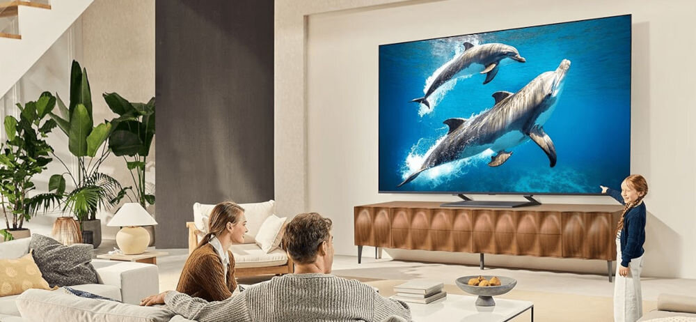 Telewizor LG 85QNED93A6A ultraduży ekran wysokiej rozdzielczości, osoby siedzące na kanapie, dziecko wskazujące na telewizor stojący na komodzie