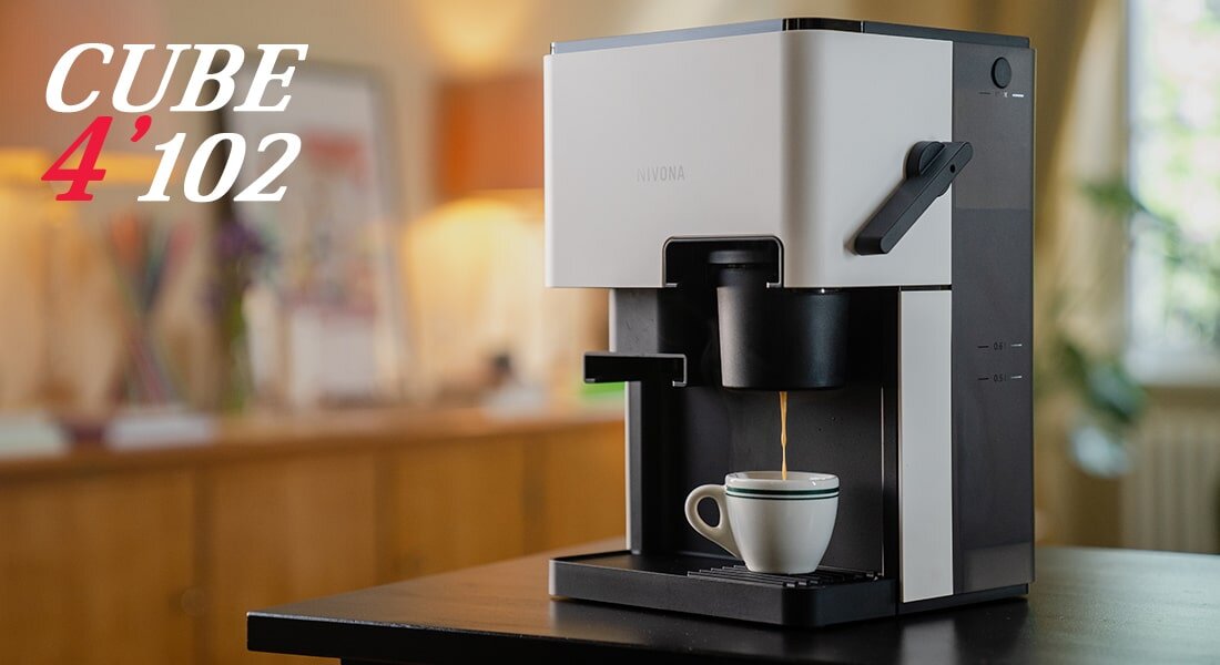 Na obrazie widoczny jest biało-czarny ekspres do kawy z filiżanką pod wylewką. W tle znajduje się rozmyte wnętrze, a na obrazie widnieje tekst: \'CUBE 4\'102\'.