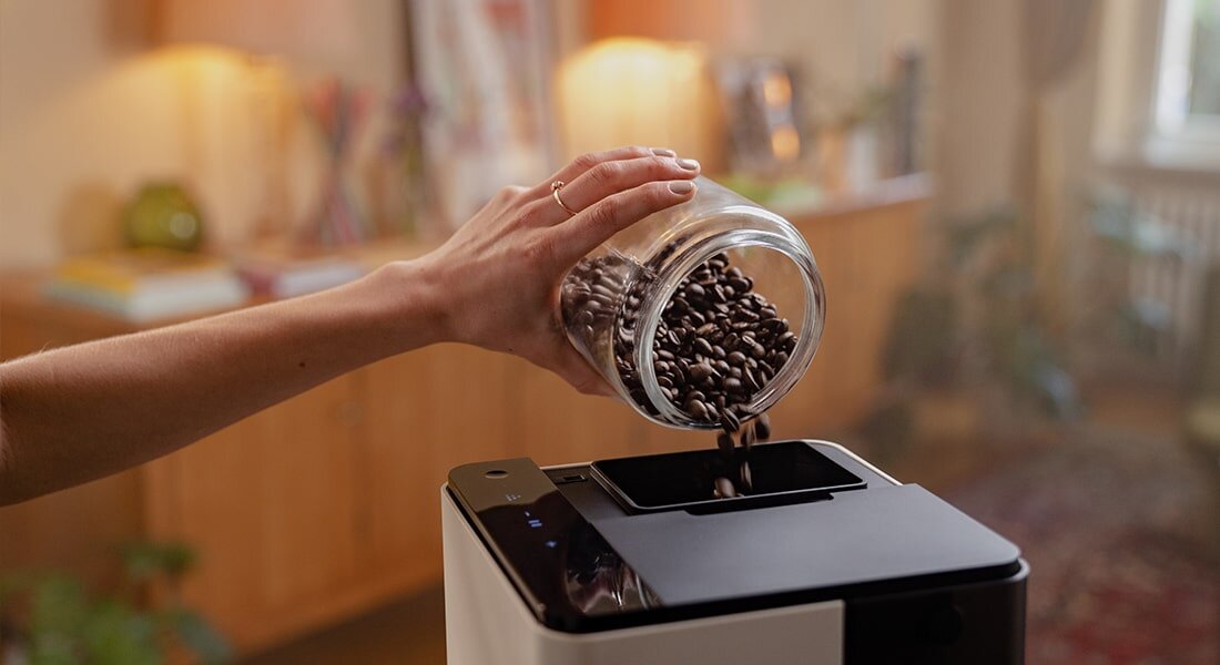 Osoba wsypuje ziarna kawy z przezroczystego słoika do ekspresu do kawy o biało-czarnym wykończeniu. W tle widoczne są rozmyte elementy wnętrza mieszkania.