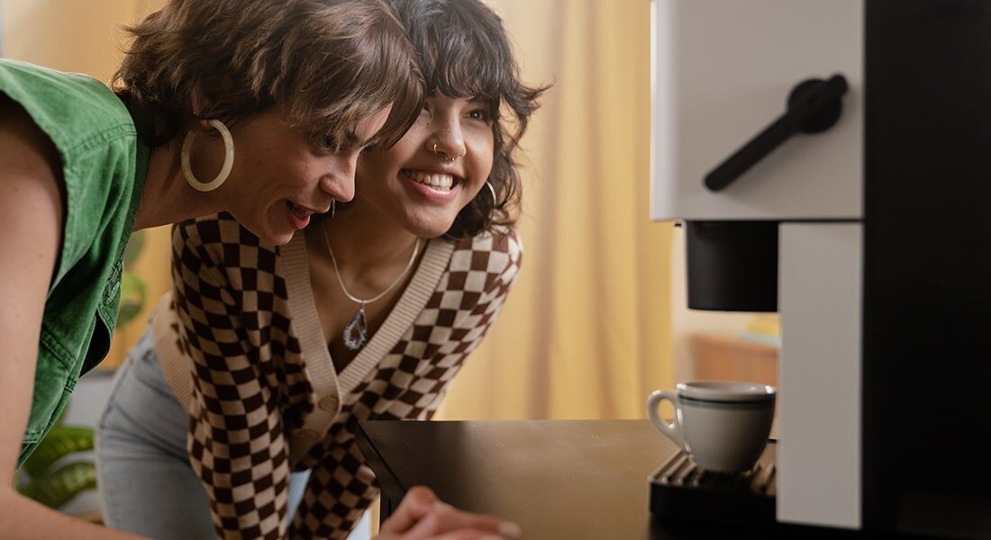 Dwie osoby patrzą na biało-czarny ekspres do kawy, na którym stoi filiżanka z kawą. Ekspres ma prosty, nowoczesny design.