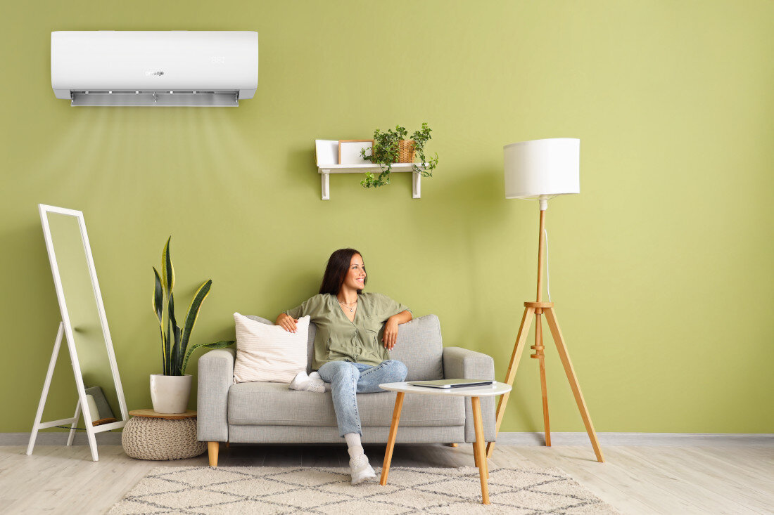 Na zielonej ścianie wisi biały klimatyzator z logo Gorenje. Przed nim znajduje się szara sofa z poduszką, na której siedzi osoba, obok stoi lampa i roślina w doniczce.