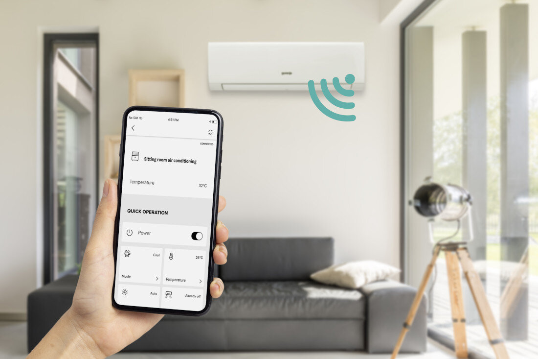 Wnętrze pokoju z zamontowanym na ścianie klimatyzatorem marki Gorenje. Osoba trzyma smartfon z aplikacją do sterowania klimatyzatorem, na ekranie widoczny jest tekst: \'Sitting room air conditioning\', \'Temperature 23°C\', \'QUICK OPERATION\', \'Power\'.