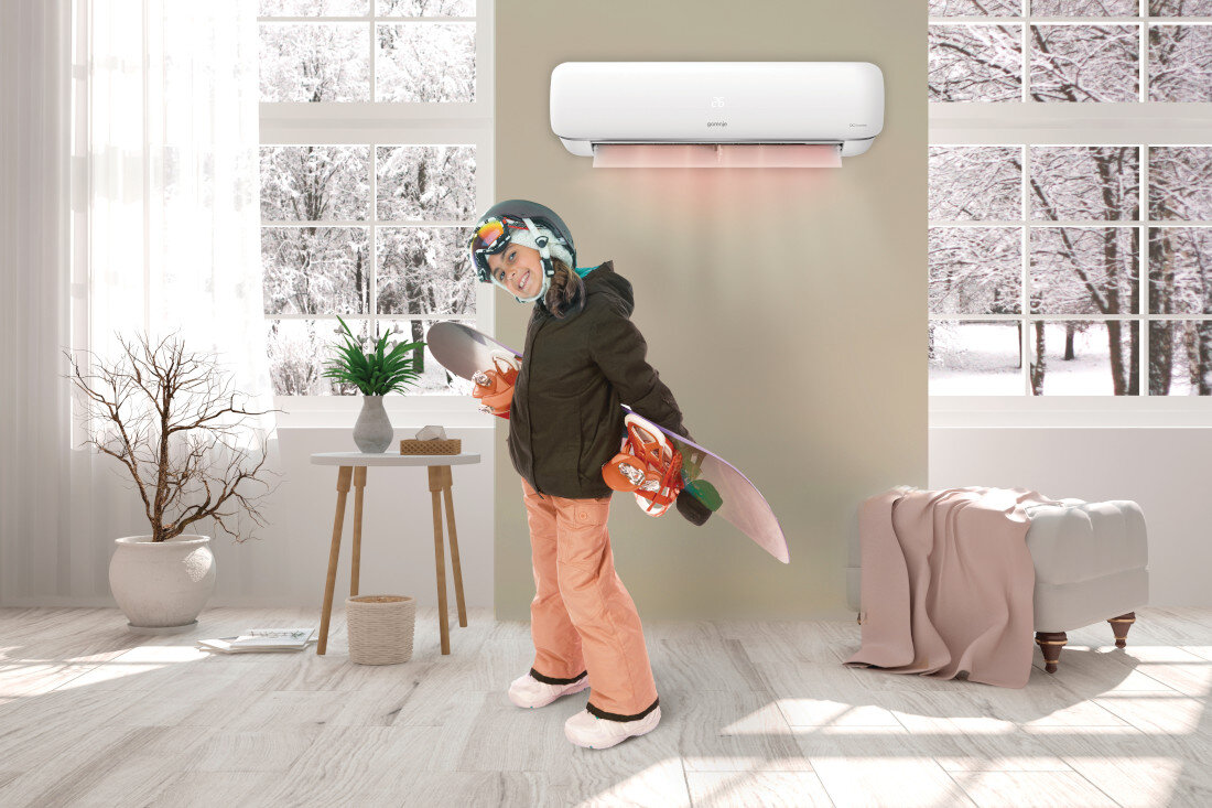 W jasnym pokoju z dużymi oknami widać klimatyzator na ścianie z logo Gorenje. Na podłodze stoi osoba w stroju zimowym z deską snowboardową.