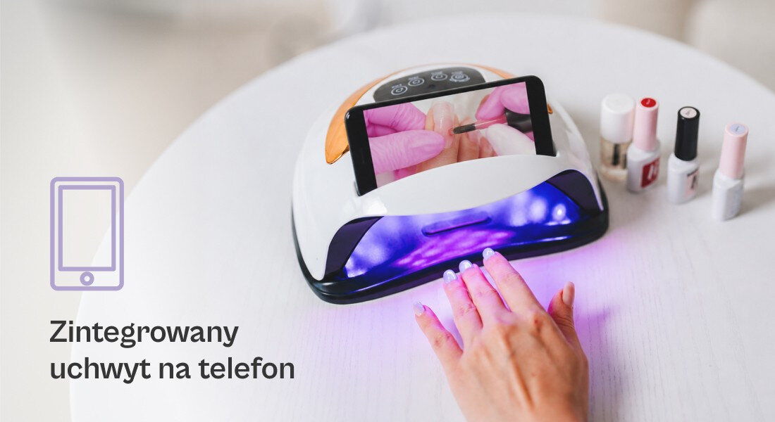Lampa UV/LED do paznokci z wbudowanym uchwytem na telefon, emitująca fioletowe światło. Obok znajdują się buteleczki z lakierami do paznokci, a na ekranie telefonu widoczny jest obraz dłoni z pomalowanymi paznokciami.