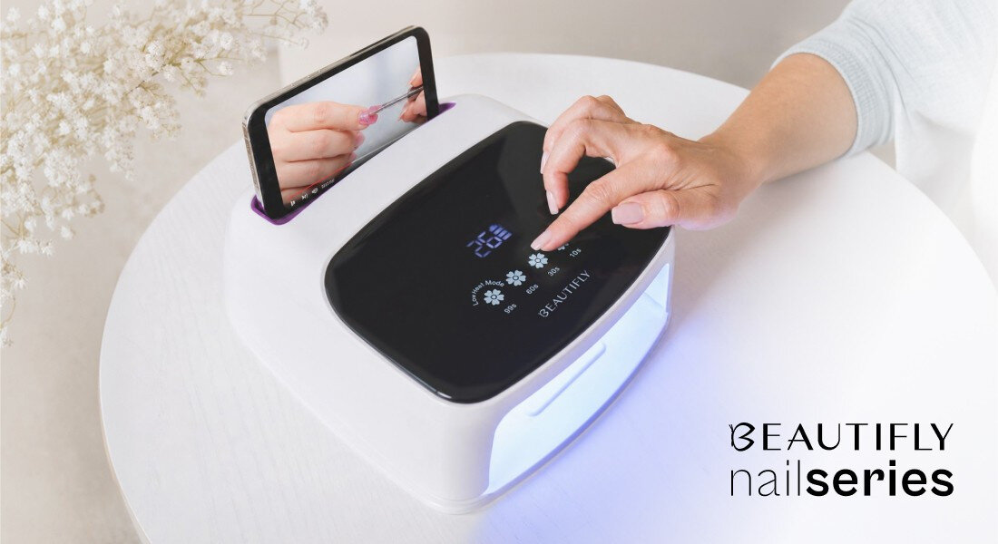 Osoba obsługuje lampę UV/LED do paznokci z wyświetlaczem i panelem dotykowym. Obok widoczny jest telefon z wyświetlanym obrazem dłoni, a w rogu napis \'BEAUTIFLY nail series\'.