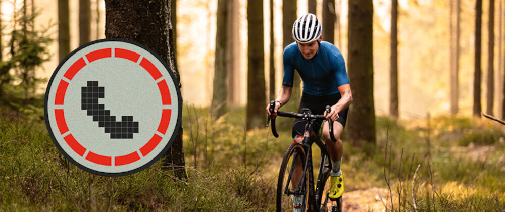 Osoba na rowerze jedzie przez las, ubrana w niebieską koszulkę i kask. Obok widoczna jest ikona telefonu w czerwonym okręgu.