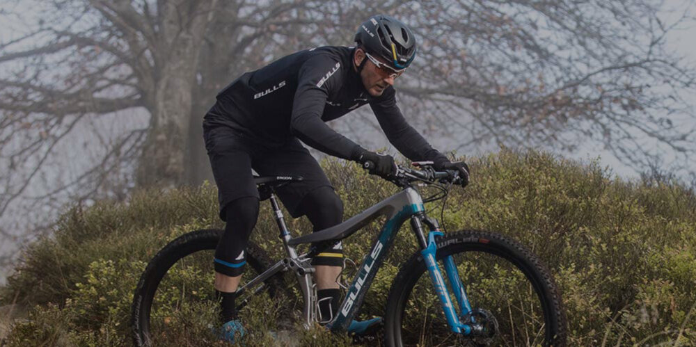 Osoba na rowerze górskim w terenie zalesionym, ubrana w kask i odzież sportową z widocznym napisem \'BULLS\'.