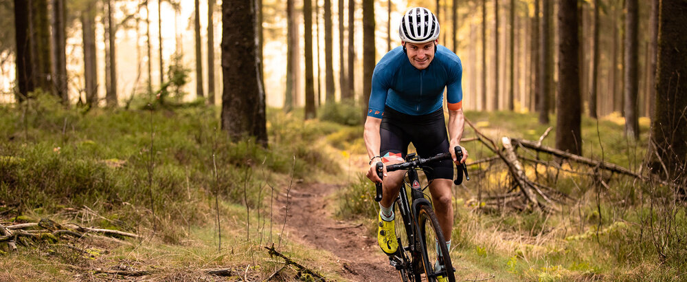 Osoba na rowerze jedzie ścieżką w lesie, ubrana w niebieską koszulkę i kask. W tle widać drzewa i zarośla.