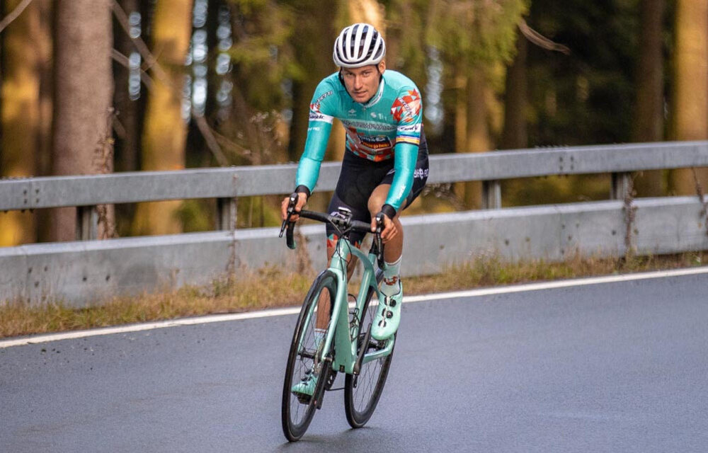 Osoba na rowerze szosowym jedzie po asfaltowej drodze, ubrana w kask i strój kolarski. W tle widać barierkę ochronną i drzewa.