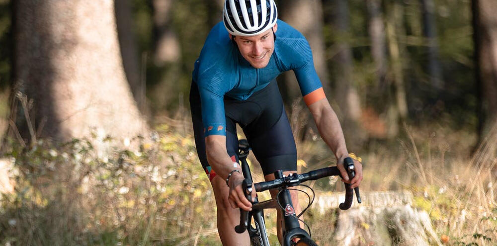 Osoba na rowerze górskim jedzie przez las, ubrana w niebieską koszulkę i kask w paski. W tle widać drzewa i trawę.