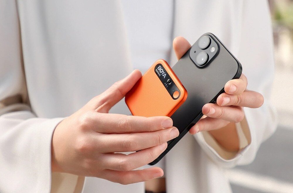 Dyktafon AI BOYA Notra Dwie dłonie trzymają czarny smartfon z przyczepionym do jego obudowy małym, pomarańczowym urządzeniem o zaokrąglonych rogach, redukcja szumów, transkrypcja