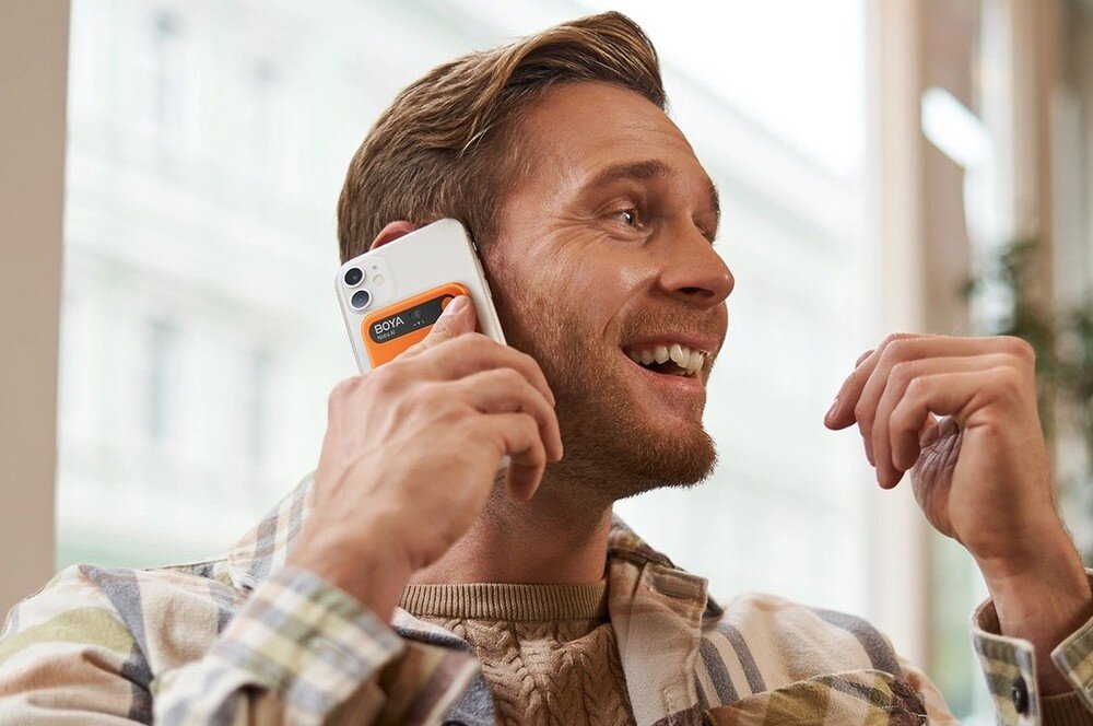 Dyktafon AI BOYA Notra Uśmiechnięty mężczyzna rozmawia przez biały smartfon, trzymając go przy uchu, a na obudowie telefonu widoczne jest małe, pomarańczowe urządzenie z napisem 'BOYA Notra AI', wbudowana pamięć, bateria, czas działania