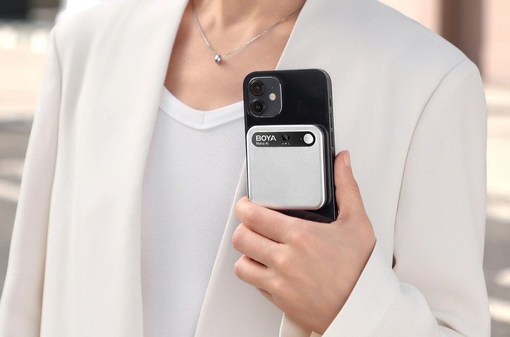Dyktafon AI BOYA Notra Bliski kadr na dłoń kobiety w białej marynarce trzymającą czarny smartfon, do którego tyłu przyczepione jest prostokątne, srebrno-czarne urządzenie z napisem 'BOYA Notra AI', opis