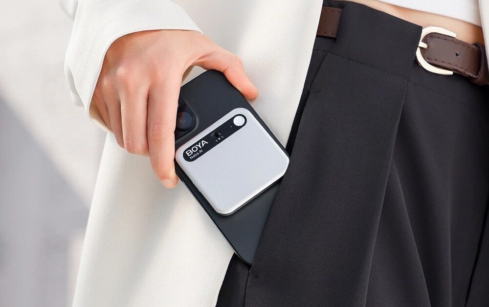 Dyktafon AI BOYA Notra Kadr na dłoń wsuwającą czarny smartfon do kieszeni ciemnych spodni, z widocznym na obudowie telefonu magnetycznym, srebrnym akcesorium o zaokrąglonych rogach, sztuczna inteligencja, pakiety, integracja z AI