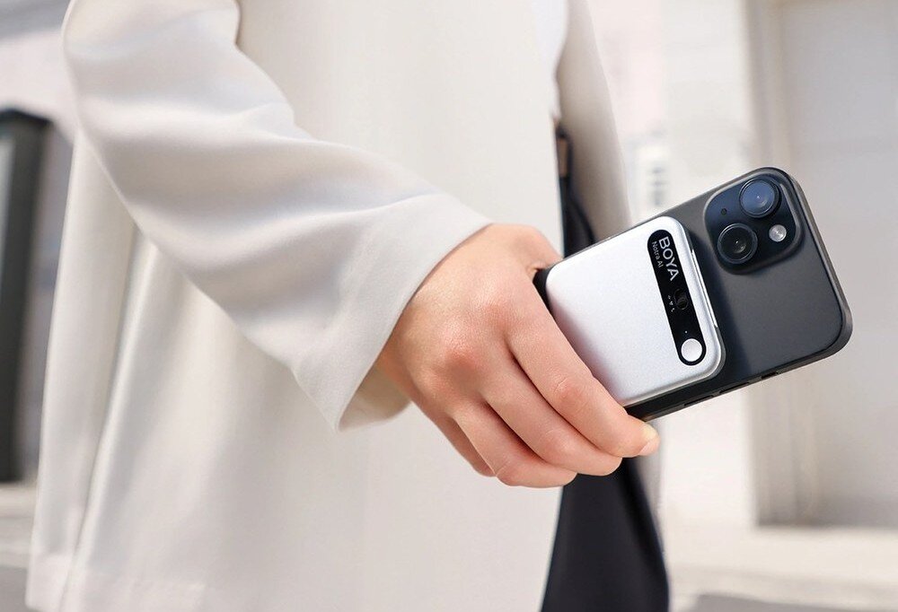 Dyktafon AI BOYA Notra Kobieta w białym żakiecie trzyma w opuszczonej dłoni czarny telefon komórkowy, do którego plecków dociśnięte jest magnetyczne, metaliczne akcesorium z czarnym paskiem i okrągłym elementem, obudowa, wymiary, kolor