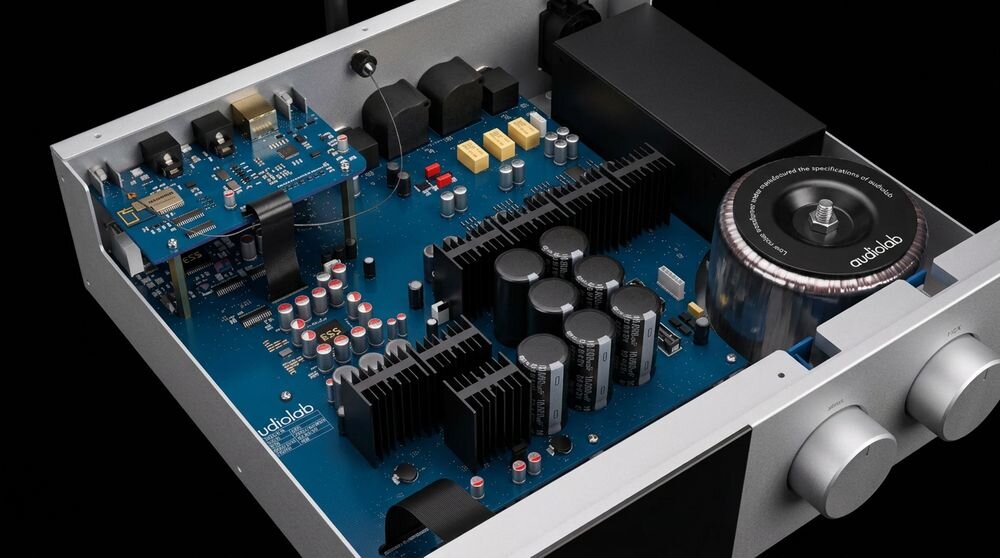 Wnętrze urządzenia elektronicznego z widocznymi komponentami na niebieskiej płytce drukowanej, w tym kondensatorami i radiatorami. Na jednym z elementów widoczne jest logo \'audiolab\'.