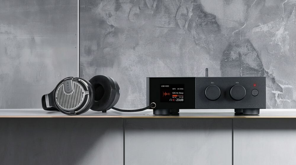 Na obrazie znajduje się czarny przetwornik C/A z dwoma pokrętłami i wyświetlaczem, na którym widoczny jest tekst \'USB IN 44.1KHZ\' oraz \'Sine Sweep\'. Obok leżą słuchawki z dużymi nausznikami i widocznym logo \'AUDIOLAB\'.