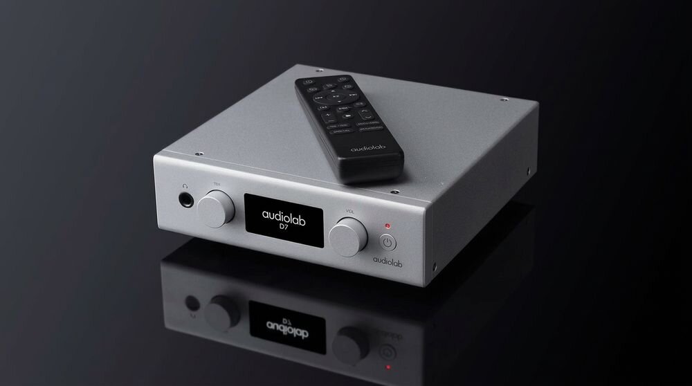 Na obrazie znajduje się srebrny przetwornik C/A z wyświetlaczem pokazującym napis \'audiolab D7\'. Na urządzeniu leży czarny pilot z widocznym logo \'audiolab\'.