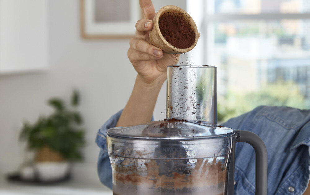 Osoba wsypuje proszek z małego naczynia do otworu robota kuchennego. W tle widać rozmyty fragment wnętrza z rośliną.