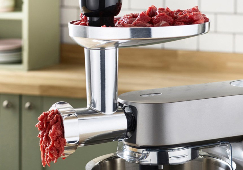 Na obrazie widoczny jest robot kuchenny z przystawką do mielenia mięsa, z którego wychodzi zmielone mięso. W tle znajduje się kuchnia z szafkami i blatem.
