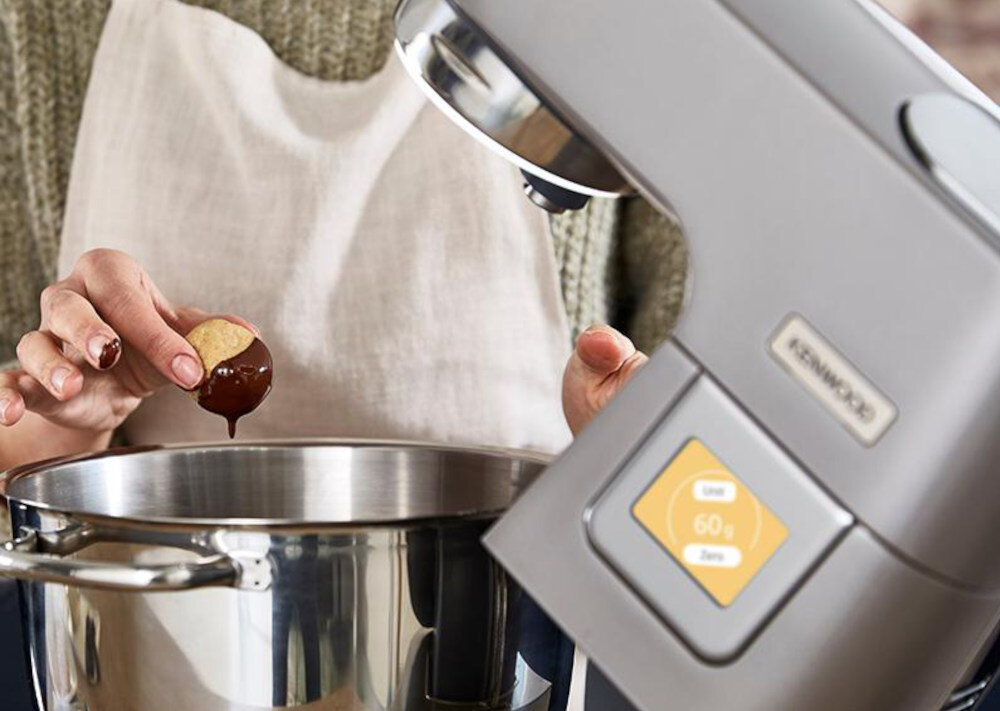 Osoba trzymająca kulkę ciasta nad dużą metalową misą robota kuchennego. Na urządzeniu widoczne jest logo \'KENWOOD\' oraz wyświetlacz z napisem \'60 g\'.
