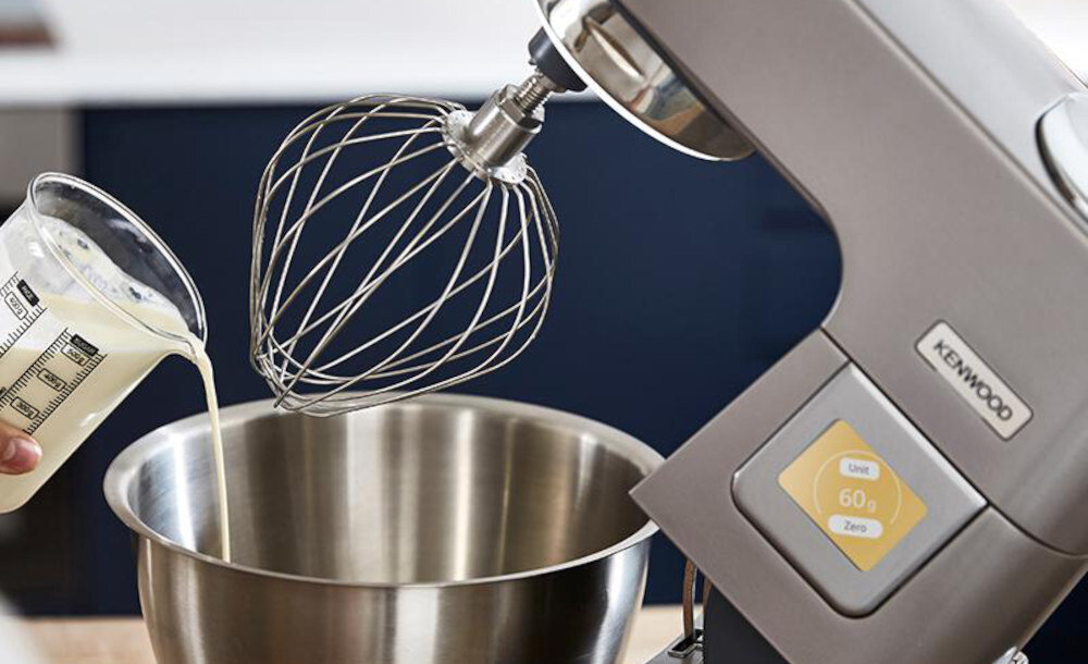 Na obrazie widoczny jest robot kuchenny z metalową misą i trzepaczką, do której wlewane jest mleko z miarki. Na urządzeniu znajduje się logo \'KENWOOD\' oraz wyświetlacz z napisem \'Unit 60 g Zero\'.