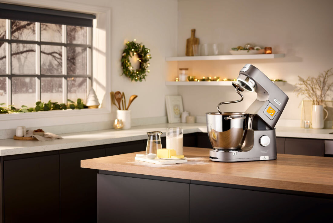 Na blacie kuchennym stoi robot kuchenny z misą i hakiem do mieszania. W tle widać okno z dekoracjami oraz półki z naczyniami i ozdobami.