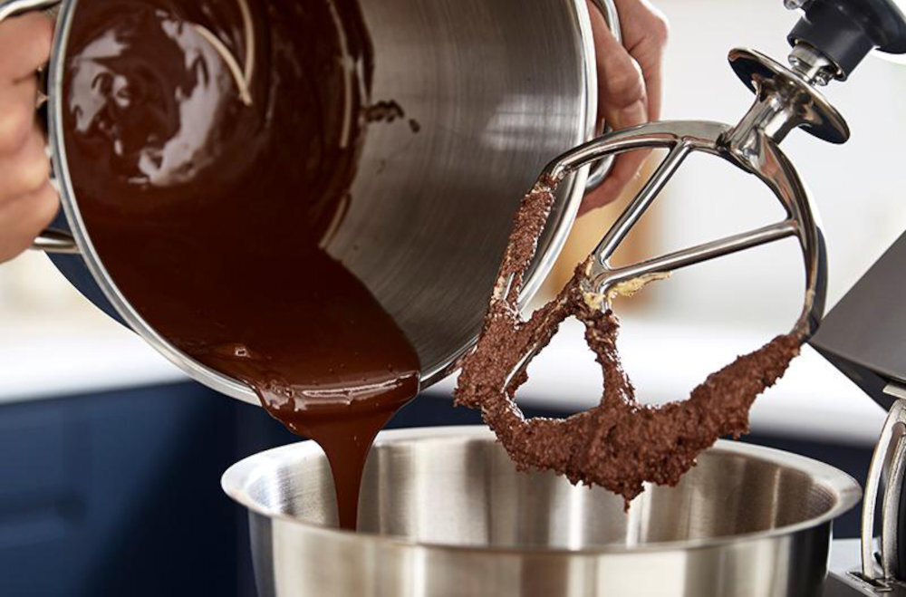 Na obrazie widać ręce trzymające metalową miskę, z której wylewa się płynna czekolada do innej miski robota kuchennego. Obok znajduje się mieszadło z resztkami czekolady.