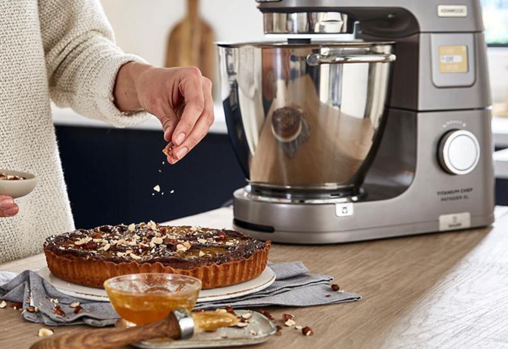 Osoba posypuje orzechami ciasto znajdujące się na drewnianym blacie, obok stoi robot kuchenny z metalową misą. Na robocie widoczne jest logo \'KENWOOD\' oraz napis \'TITANIUM CHEF PATISSIER XL\'.