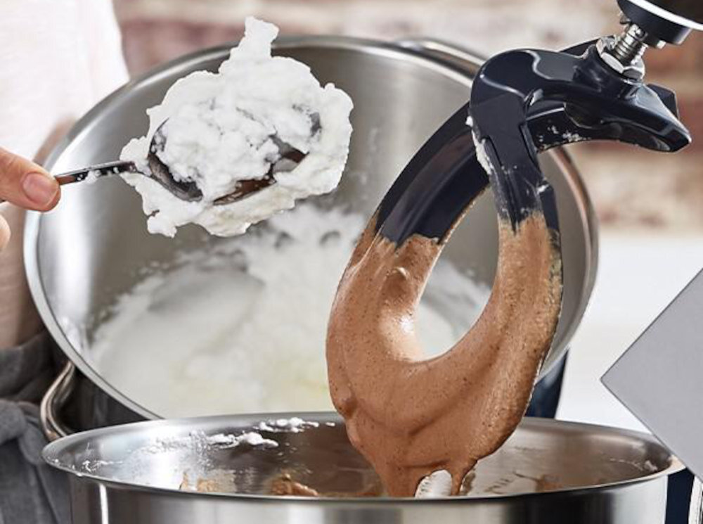 Na obrazie widać robot kuchenny z mieszadłem, które miesza brązową masę w metalowej misie. Obok znajduje się łyżka z białą masą, trzymana nad drugą misą.