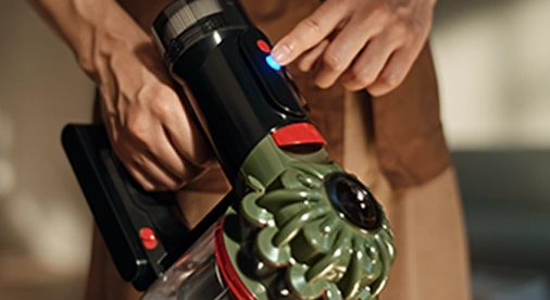 Osoba trzyma odkurzacz z widocznym przyciskiem i niebieskim wskaźnikiem świetlnym. Odkurzacz ma zielony element z przodu i przezroczysty pojemnik na kurz.