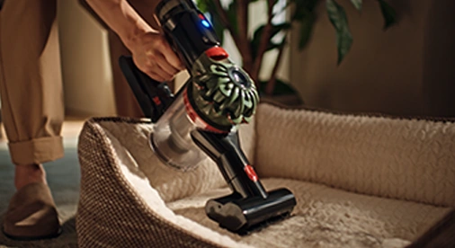 Osoba używa odkurzacza ręcznego do czyszczenia legowiska dla zwierząt. W tle widoczne są rośliny doniczkowe.