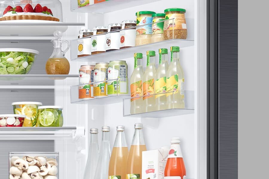 Wnętrze lodówki z półkami wypełnionymi różnorodnymi produktami spożywczymi, takimi jak napoje w butelkach i puszkach, słoiki z przetworami oraz pojemniki z sałatkami. Na drzwiach lodówki znajdują się dodatkowe półki z napojami i słoikami.