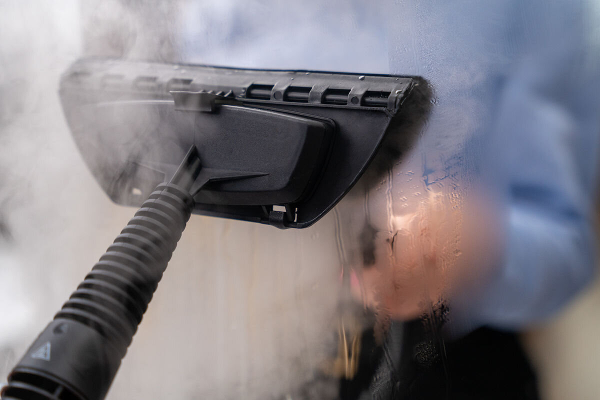 Na obrazie widoczny jest czyścik parowy używany do czyszczenia zaparowanej powierzchni. W tle widać rozmytą postać w niebieskiej koszuli.