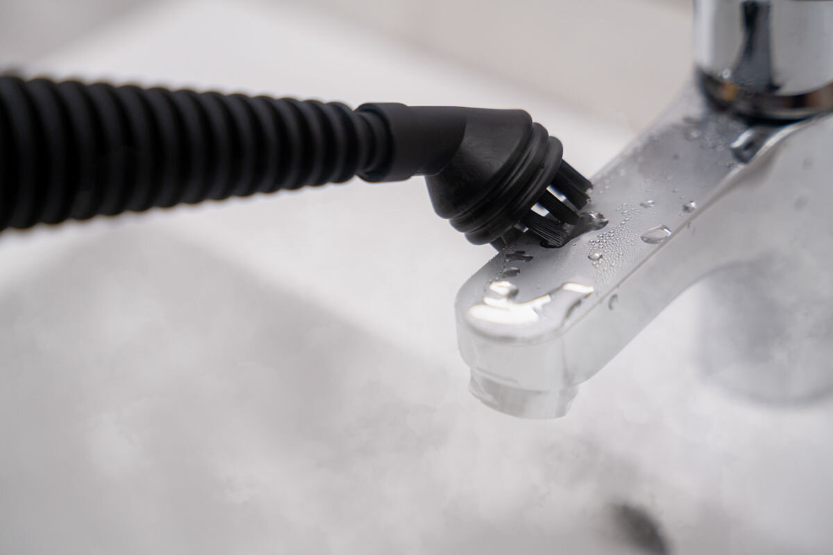 Czarny czyścik parowy z wąską końcówką skierowany na metalową baterię łazienkową. Na powierzchni baterii widoczne są krople wody.