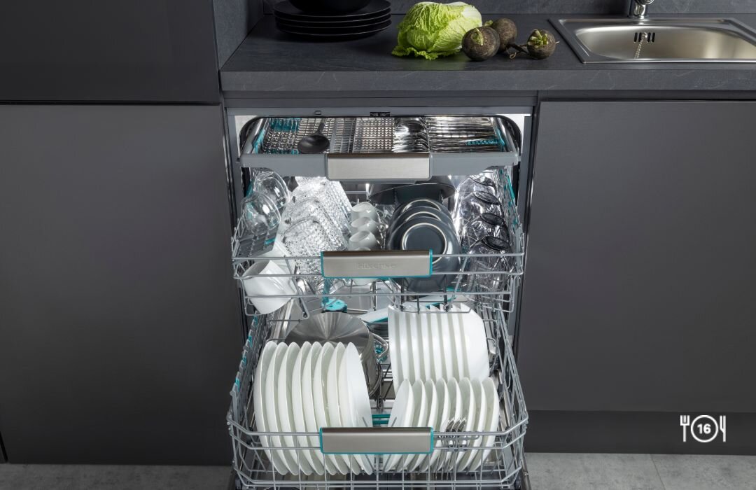 Otwarta zmywarka z widocznymi dwoma koszami pełnymi naczyń, w tym talerzy i szklanek. Na blacie obok zlewu znajdują się warzywa, w tym sałata i awokado.