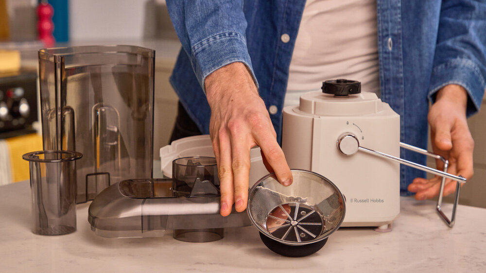 Osoba składa sokowirówkę, trzymając metalowy element nad blatem. Na urządzeniu widoczne jest logo \'Russell Hobbs\'.