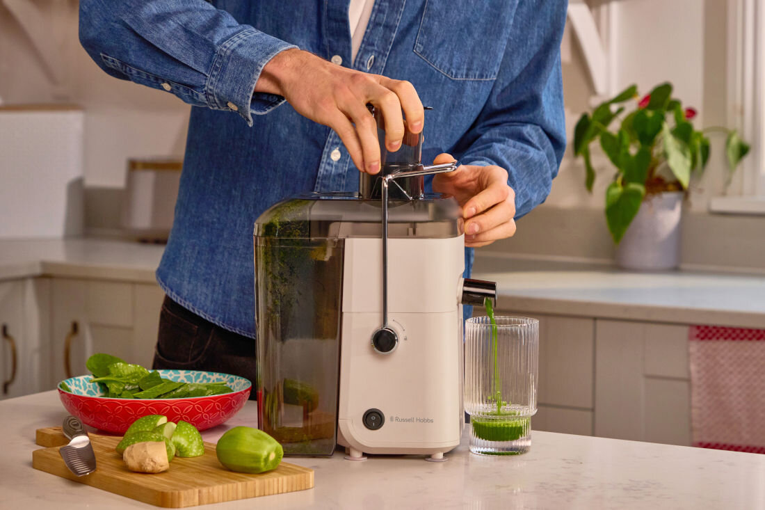 Osoba obsługuje beżową sokowirówkę marki Russell Hobbs, z której do szklanki nalewany jest zielony sok. Na blacie obok znajdują się pokrojone owoce i warzywa oraz miska z zielonymi liśćmi.
