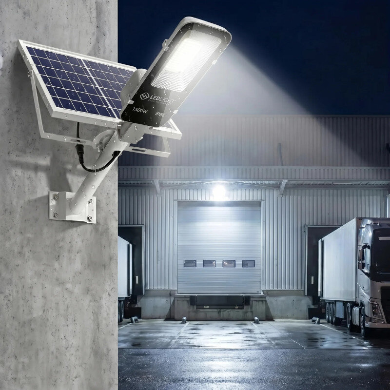 Na ścianie zamontowana jest lampa solarna LED z panelem słonecznym, emitująca jasne światło. W tle widoczny jest budynek przemysłowy z zaparkowanymi ciężarówkami.