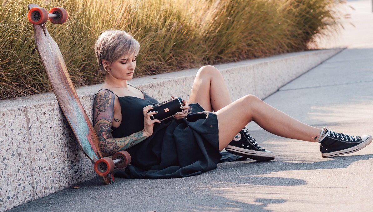 Osoba siedzi na chodniku obok deskorolki, trzymając w rękach czarny głośnik mobilny. W tle widoczne są trawy ozdobne.