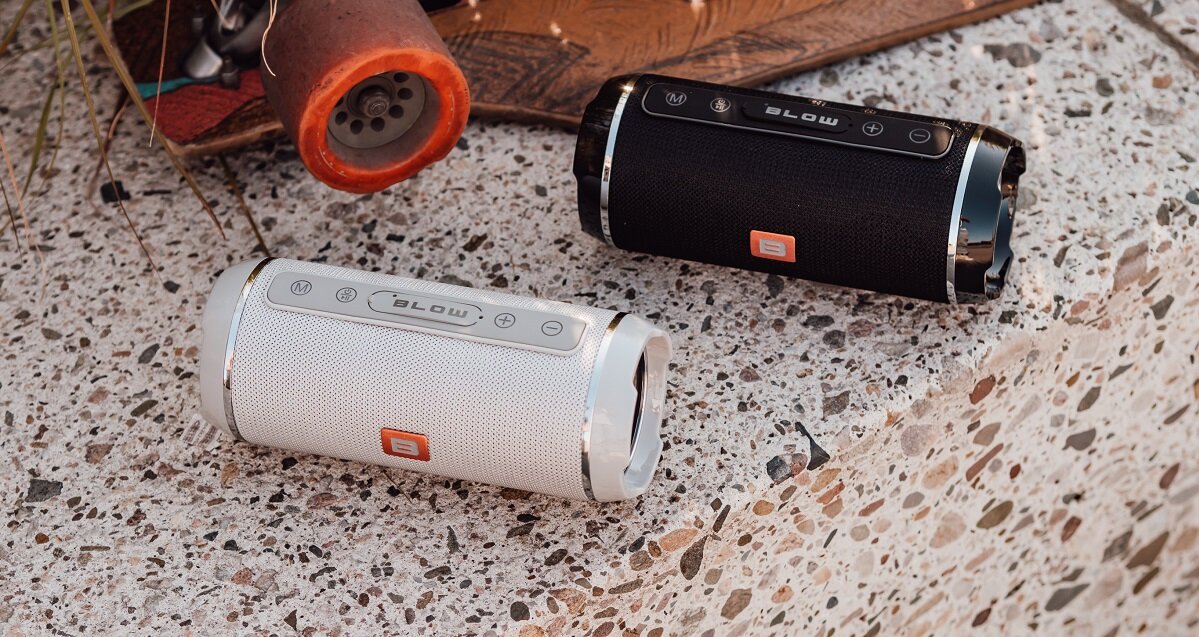 Na obrazie znajdują się dwa głośniki mobilne marki BLOW, jeden w kolorze czarnym, drugi w białym, umieszczone na powierzchni z wzorem przypominającym terrazzo. Na głośnikach widoczne są przyciski sterujące oraz logo BLOW.