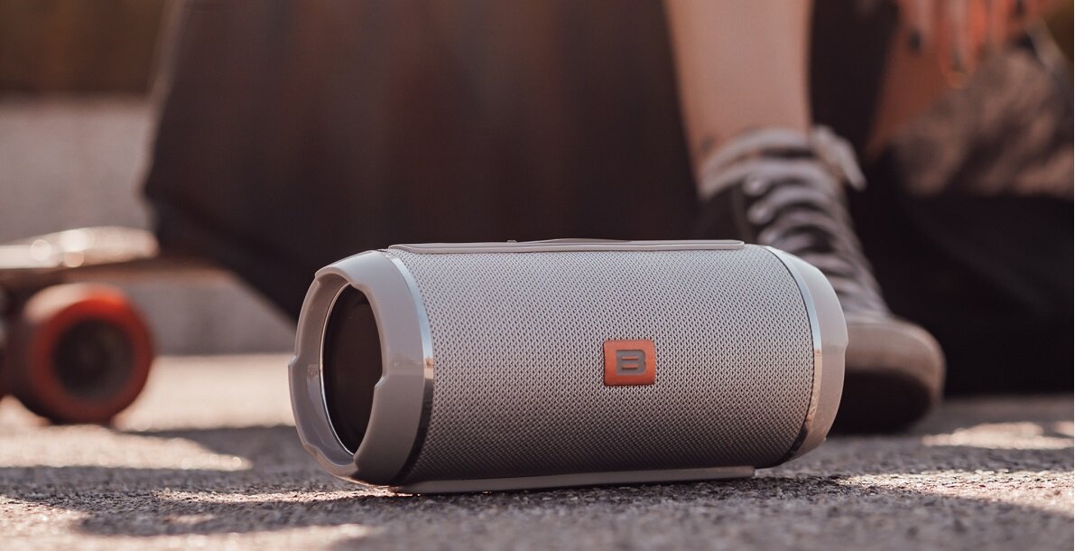 Na obrazie widoczny jest przenośny głośnik o cylindrycznym kształcie z metaliczną siatką i logo w kształcie litery B. W tle znajduje się deskorolka i osoba w sportowym obuwiu.