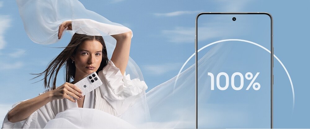Osoba trzymająca biały smartfon na tle nieba z chmurami. Obok widoczny jest ekran telefonu z napisem \'100%\'.