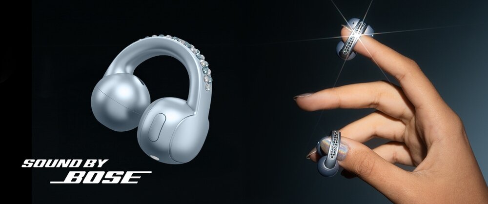 Na obrazie widoczne są srebrne słuchawki o nowoczesnym designie, trzymane przez dłoń. Obok znajduje się napis \'SOUND BY BOSE\'.