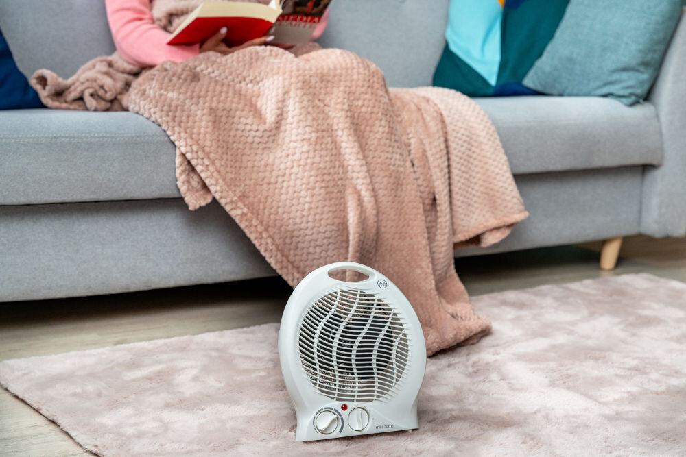 Na dywanie przed sofą stoi biały termowentylator z dwoma pokrętłami. Na sofie siedzi osoba przykryta kocem, trzymająca książkę.