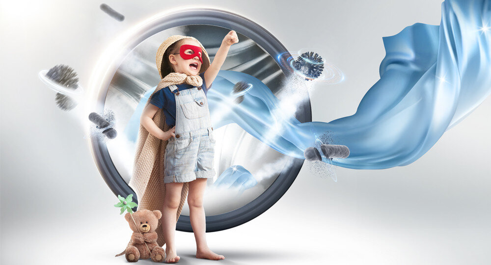 Dziecko w czerwonej masce i pelerynie stoi przed dużym bębnem pralki, unosząc rękę w geście triumfu. Obok znajduje się pluszowy miś, a wokół wirują niebieskie tkaniny i pędzle.