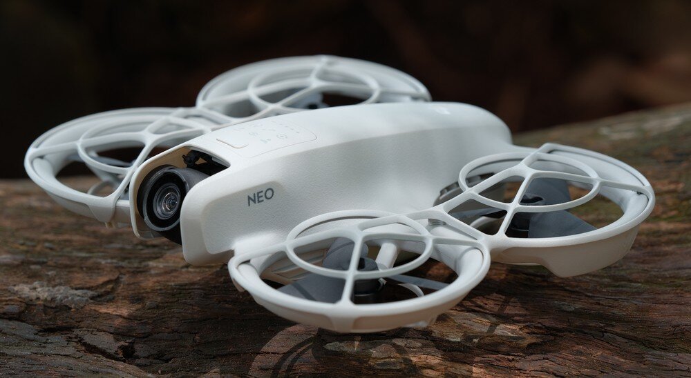 Biały dron z napisem \'NEO\' na boku, wyposażony w kamerę i osłony na śmigła, umieszczony na drewnianej powierzchni.